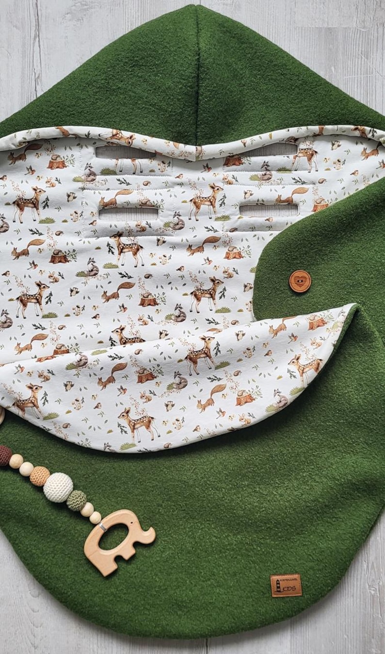 May include: A green baby stroller blanket with a hood and a woodland animal print lining. The blanket features a button closure and a wooden elephant-shaped teether with a beaded string. The lining displays deer, foxes, and other forest creatures.