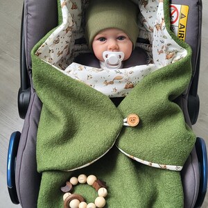 May include: A baby wrapped in a green hooded blanket with a woodland animal print lining, secured with a wooden button. The baby is wearing a matching green hat and has a pacifier. A wooden teether is attached to the blanket.