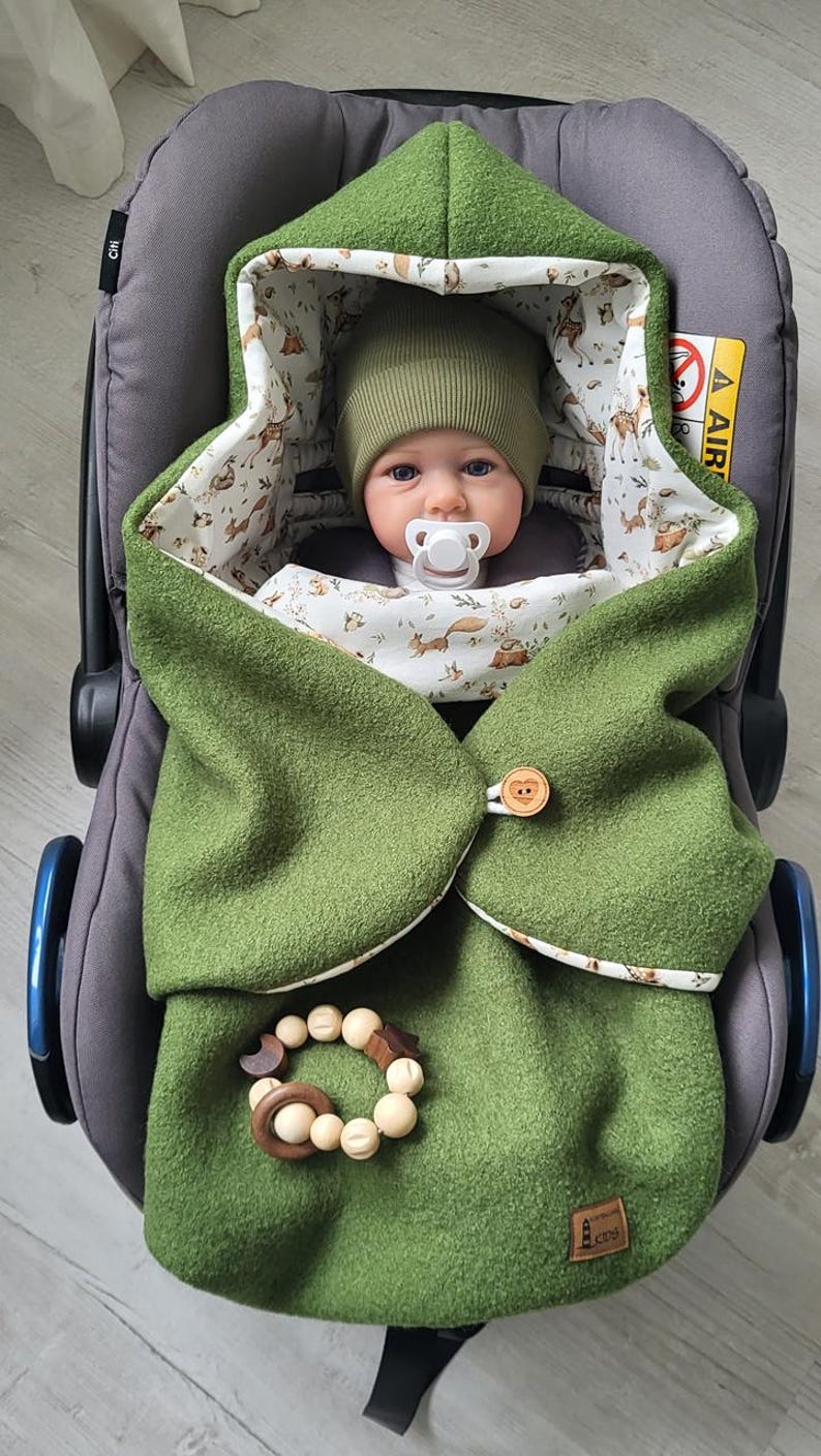 May include: A baby wrapped in a green hooded blanket with a woodland animal print lining, secured with a wooden button. The baby is wearing a matching green hat and has a pacifier. A wooden teether is attached to the blanket.