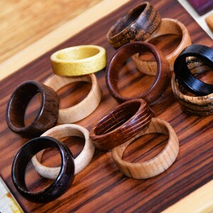 May include: A collection of wooden rings in various shades of brown, black, and yellow. The rings are stacked and arranged on a wooden surface.