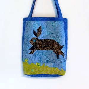 Puede incluir: Una bolsa de mano azul con una aplicación de conejo marrón sobre un fondo estampado azul y verde.