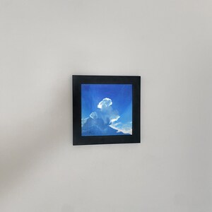May include: A framed painting of a blue sky with a large white cloud. The painting is in a black frame and is hanging on a white wall.