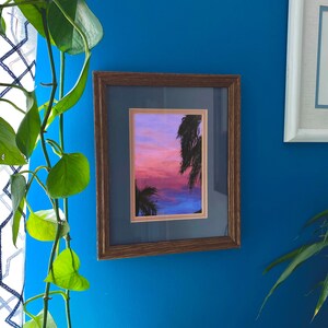 Könnte beinhalten: Ein gerahmtes Gemälde eines Sonnenuntergangs über einem Gewässer mit Palmen, die sich vor dem Himmel abzeichnen. Das Gemälde ist in einem braunen Rahmen mit Passepartout.