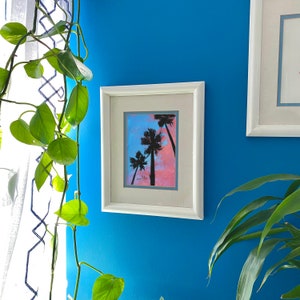 Puede incluir: Una pintura enmarcada en blanco que representa una puesta de sol rosa y azul con tres palmeras silueteados contra el cielo.