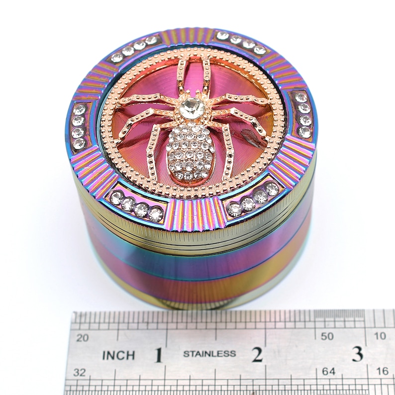 May include: A multi-colored, round grinder with a gold spider design on top, encrusted with rhinestones. The grinder has a rainbow finish and is accented with additional rhinestones around the edges. A stainless steel ruler is in the foreground.