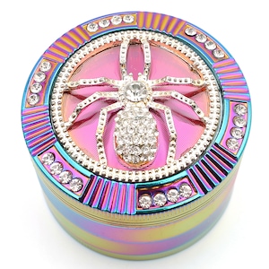 May include: A round, iridescent grinder with a bedazzled spider design. The grinder features a pink center with a gold spider encrusted with rhinestones. The outer rim is adorned with clear rhinestones and a rainbow finish.