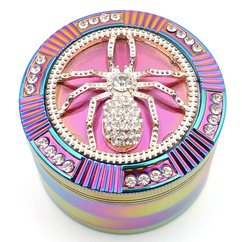 May include: A round, iridescent grinder with a bedazzled spider design. The grinder features a pink center with a gold spider encrusted with rhinestones. The outer rim is adorned with clear rhinestones and a rainbow finish.