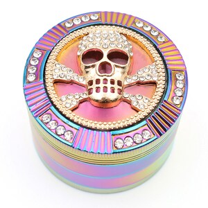 May include: A multi-colored, round grinder with a gold skull and crossbones design. The skull is encrusted with clear rhinestones, and the grinder has a rainbow finish with additional rhinestone accents.