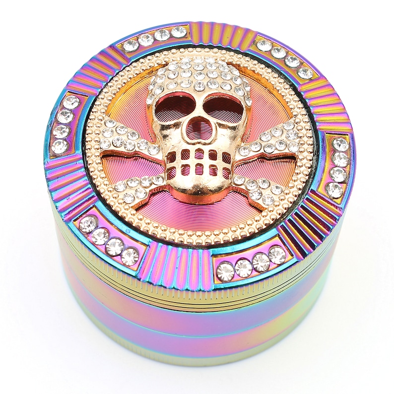 May include: A multi-colored, round grinder with a gold skull and crossbones design. The skull is encrusted with clear rhinestones, and the grinder has a rainbow finish with additional rhinestone accents.