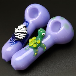 May include: Two purple glass pipes with animal figurines. One pipe has a black and white frog figurine and the other has a green turtle figurine with yellow accents.