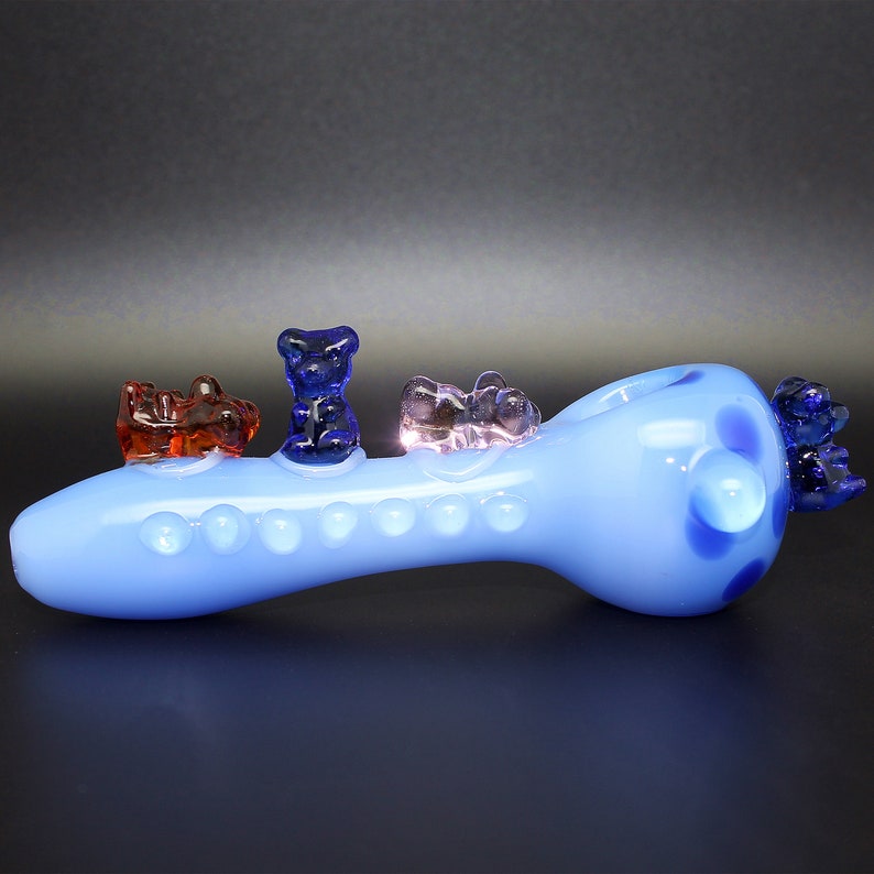 May include: A light blue glass pipe with a spotted design and four gummy bear shaped accents. The gummy bears are red, blue, and purple.