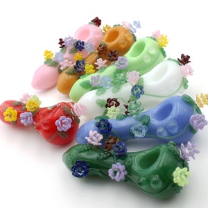 Pode incluir: Um conjunto de sete cachimbos de vidro coloridos decorados com desenhos florais. Cada cachimbo apresenta uma cor e um padrão floral diferente, incluindo rosa, verde, branco, azul, vermelho, laranja e amarelo.
