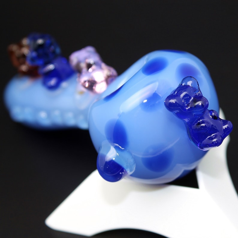 May include: A blue glass pipe with a polka dot pattern and a blue glass bear figurine attached to the side. The pipe is on a white stand.