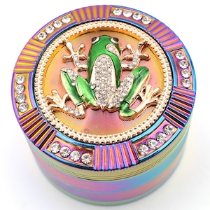 May include: A multi-colored grinder with a green frog design. The frog is embellished with rhinestones and sits on a gold-colored background. The grinder is ringed with rhinestones and has a rainbow finish.