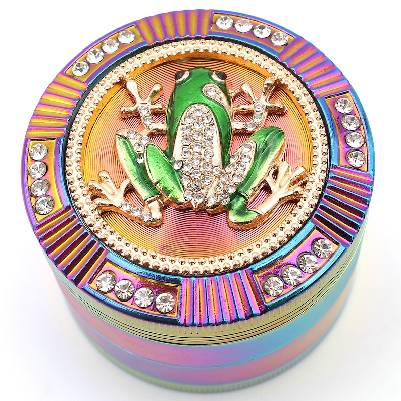 May include: A multi-colored grinder with a green frog design. The frog is embellished with rhinestones and sits on a gold-colored background. The grinder is ringed with rhinestones and has a rainbow finish.