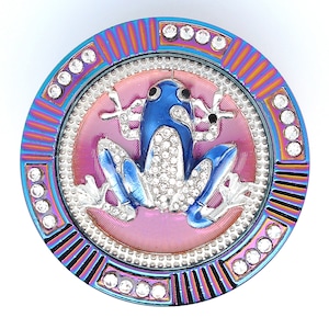 May include: A round, jeweled frog-themed trinket. The frog is blue with crystal accents, set against a pink and silver background. The outer ring is iridescent with crystal embellishments, creating a decorative and eye-catching design.