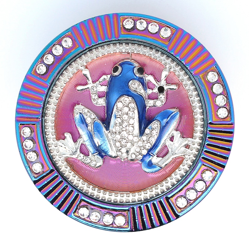May include: A round, jeweled frog-themed trinket. The frog is blue with crystal accents, set against a pink and silver background. The outer ring is iridescent with crystal embellishments, creating a decorative and eye-catching design.