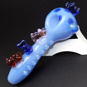 May include: A light blue glass pipe with a round bowl and a long stem. The pipe is decorated with five small glass bears in different colors, including blue, pink, and brown.