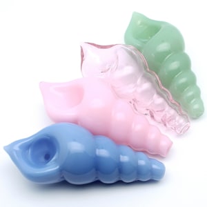 May include: Four glass seashell pipes in different colours: light blue, pink, light pink, and green. The pipes are all shaped like seashells and have a small bowl at the top for smoking.