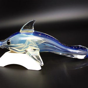 May include: A glass dolphin sculpture with a blue and white iridescent finish. The dolphin is posed in a swimming position with its tail raised.