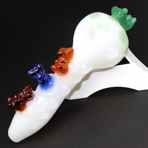 May include: White glass pipe with a green, blue, orange, and red glass bear figurine on top. The pipe is on a white stand.