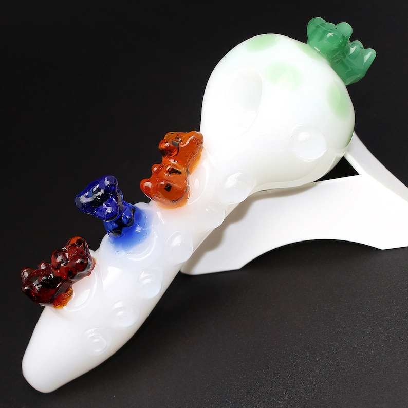 May include: White glass pipe with a green, blue, orange, and red glass bear figurine on top. The pipe is on a white stand.