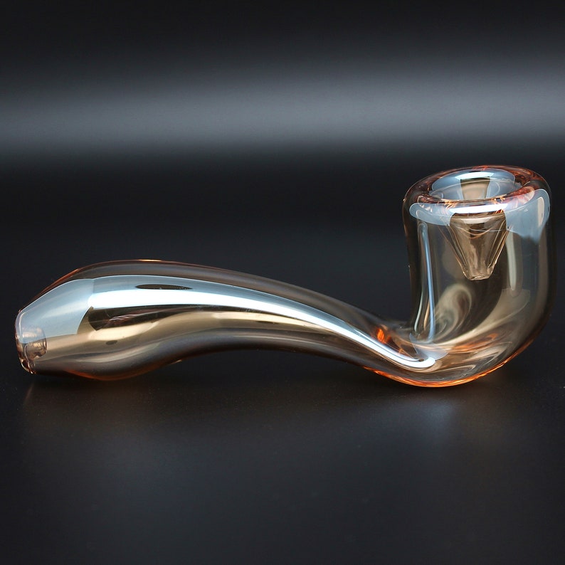 May include: A clear glass pipe with a curved stem and a bowl. The pipe has a light amber color and a reflective surface.