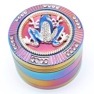 May include: A multi-colored grinder with a blue frog design. The frog is embellished with rhinestones and sits atop a pink and silver circular design. The grinder is accented with rhinestones around the edges.