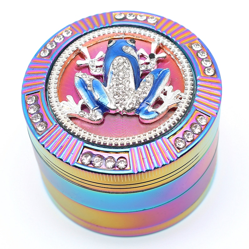May include: A multi-colored grinder with a blue frog design. The frog is embellished with rhinestones and sits atop a pink and silver circular design. The grinder is accented with rhinestones around the edges.