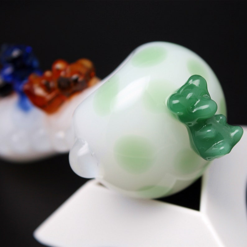 May include: White glass bead with green polka dots and a green glass animal figurine attached to the side.