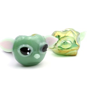 May include: Green glass pipe shaped like a cartoon character with large eyes and ears. The pipe has a small hole on the top for smoking.