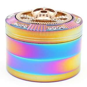 May include: A multi-colored, cylindrical grinder with a skull design on top. The grinder has a rainbow finish with gold accents and is adorned with clear crystals. The skull is gold-colored and centered on the top.