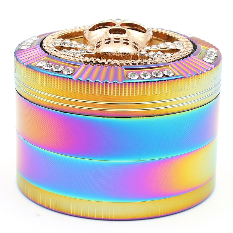 May include: A multi-colored, cylindrical grinder with a skull design on top. The grinder has a rainbow finish with gold accents and is adorned with clear crystals. The skull is gold-colored and centered on the top.