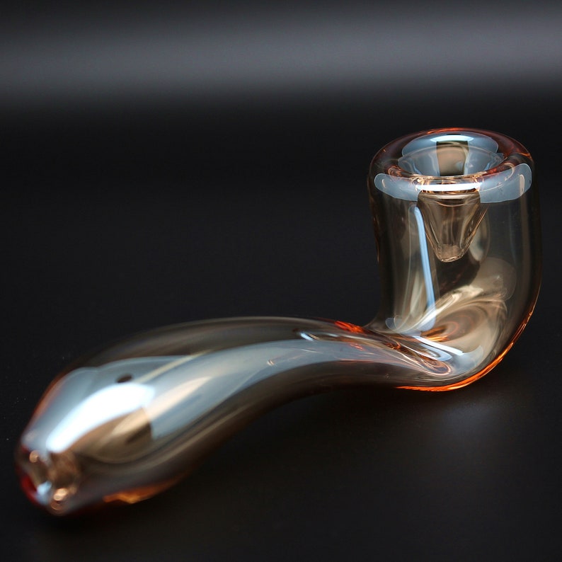 May include: A clear glass pipe with a curved design. The pipe has a rounded bowl and a long, slender stem. The glass has a slight amber tint.