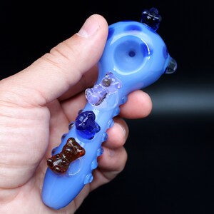 May include: A light blue glass pipe with a rounded bowl and a textured handle. The pipe is decorated with four colorful glass bears.