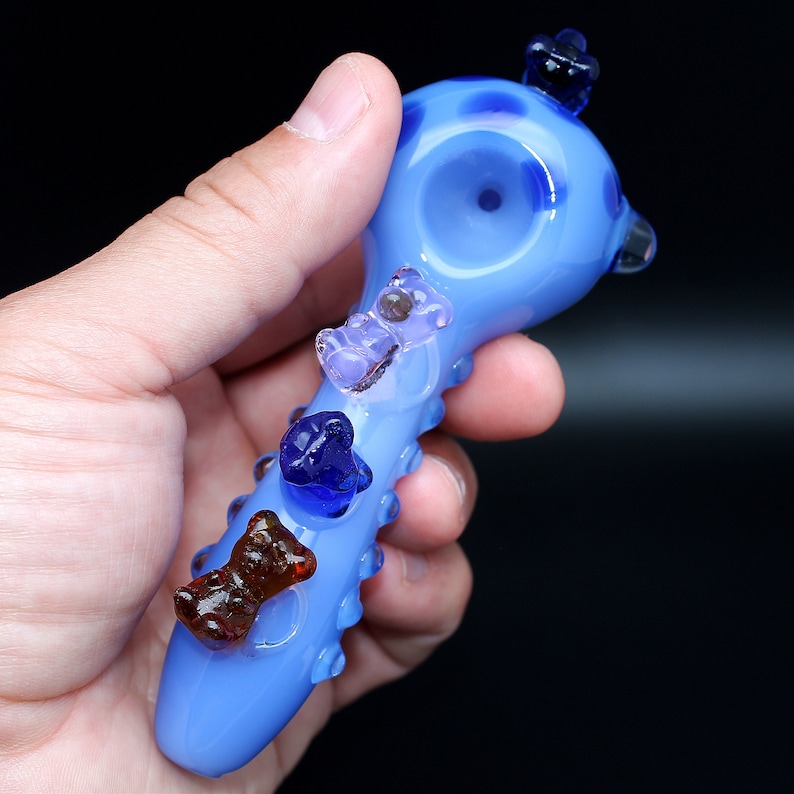 May include: A light blue glass pipe with a rounded bowl and a textured handle. The pipe is decorated with four colorful glass bears.