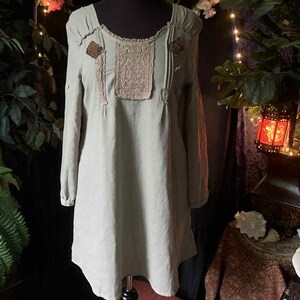 Può includere: Un abito tunica verde chiaro con maniche lunghe e scollatura quadrata. L'abito presenta toppe decorative e dettagli in pizzo sul corpetto. L'abito è esposto su un manichino nero. Lo sfondo comprende vegetazione e luci fatate.