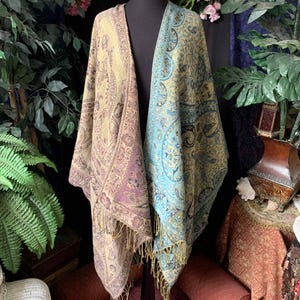 Puede incluir: Dos chales con estampado de cachemira, uno en tonos lavanda y crema, el otro en turquesa y crema, se exhiben en un maniquí. Ambos chales tienen bordes con flecos. El fondo incluye vegetación y un letrero que dice "The Witch Apparel."