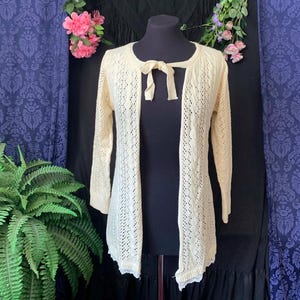 Puede incluir: Rebeca crema con diseño calado y frontal abierto, con cierre de lazo en el cuello. El suéter de manga larga se exhibe en un maniquí negro sobre un fondo de tela morada y detalles florales. La rebeca tiene un dobladillo festoneado.