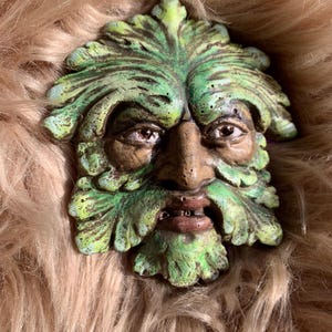 Puede incluir: Una escultura detallada de la cara de un hombre verde, con barba y corona de hojas. La cara es una mezcla de marrón y verde, con ojos detallados y una superficie texturizada. La escultura probablemente está hecha de cerámica o un material similar.