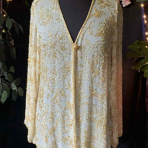 May include: A cream-coloured, long-sleeved top with gold floral beading. The top has a V-neck and a single button closure. The garment is displayed on a black mannequin against a dark background with floral accents and a sign that reads "Vintage Apparel".