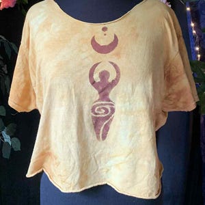 Peut inclure: Haut court jaune moutarde avec un motif bordeaux représentant une femme avec un croissant de lune et un motif en spirale. Le haut a des manches courtes et un col rond. Le haut est présenté sur un mannequin noir.