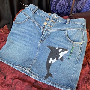 Op de afbeelding: Een blauwe denim minirok met een zwart-witte orka-walvis afbeelding en groene kralen. De rok heeft een knoopsluiting en past op een riem tot 5 cm breed.