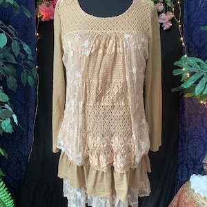 Op de afbeelding: Een beige top met lange mouwen en ingewikkelde kant- en haakdetails. De top heeft een ronde halslijn en een gelaagde, gerimpelde zoom. Witte bloemenkant siert de voorkant en voegt een delicate touch toe aan het kledingstuk.