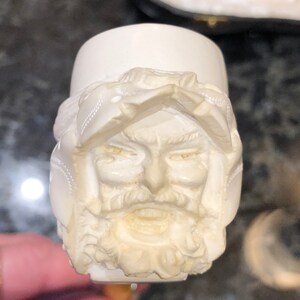 Może przedstawiać: Biała ceramiczna fajka z rzeźbą twarzy mężczyzny. Fajka jest w tradycyjnym stylu z misą i trzonkiem.