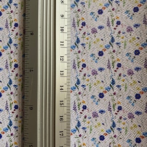 May include: A close-up of a white fabric with a small floral pattern in shades of blue, purple, yellow, and green. A silver ruler is placed on the fabric, with the numbers 6 through 19 visible.