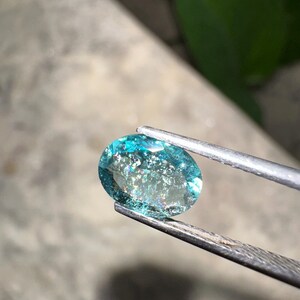 May include: An oval-cut, aquamarine gemstone held by tweezers. The gemstone is a vibrant teal color with internal inclusions that create a shimmering effect. The background is blurred, with hints of green foliage.