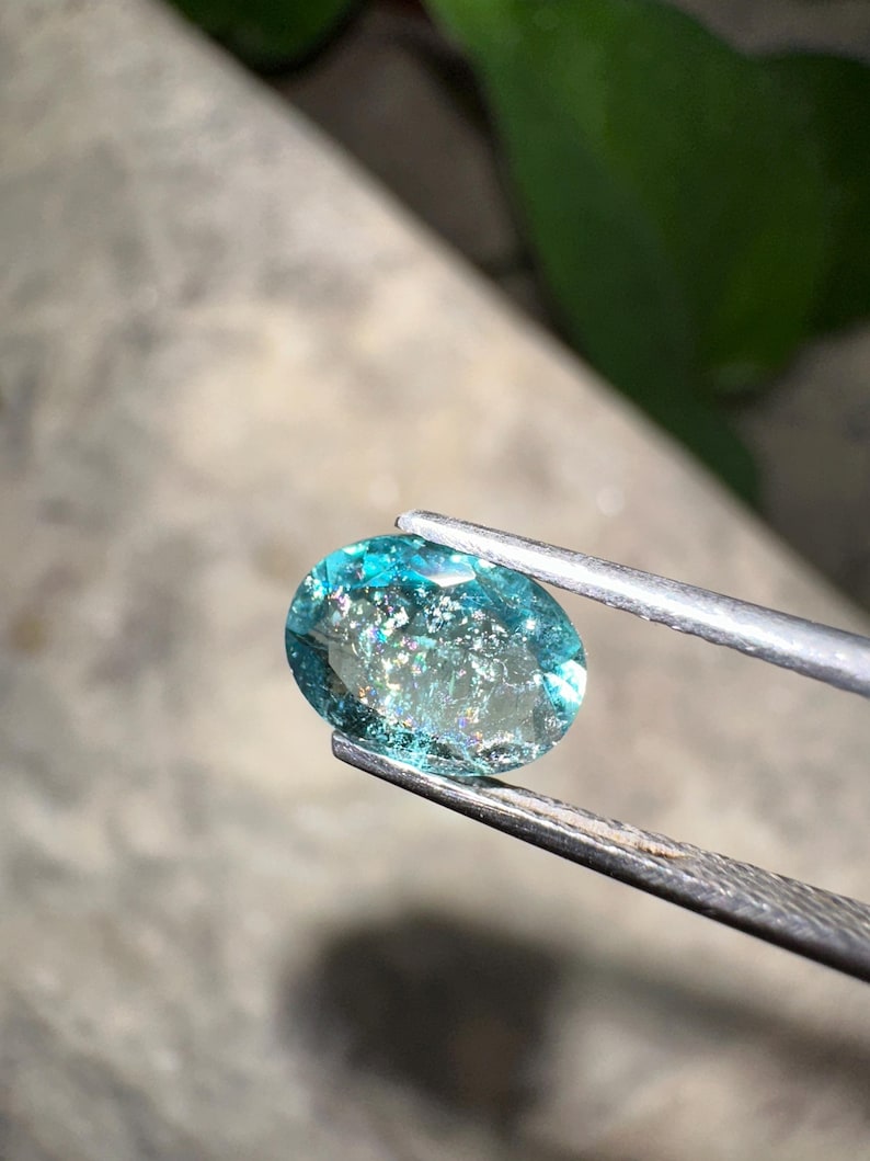 May include: An oval-cut, aquamarine gemstone held by tweezers. The gemstone is a vibrant teal color with internal inclusions that create a shimmering effect. The background is blurred, with hints of green foliage.