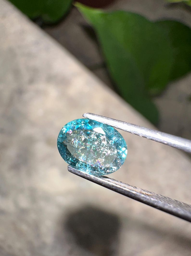 May include: An oval-shaped, teal gemstone held by tweezers. The gem has internal inclusions and a shimmering, iridescent quality. The background is blurred, with hints of green foliage and a gray surface.