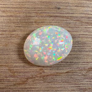 May include: Oval-shaped opal gemstone with a milky white base and iridescent flashes of color. The opal displays a play-of-color effect, with vibrant hues of red, green, and blue. The gemstone is set against a wooden surface.